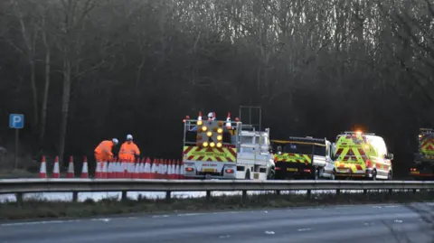 RSM Photography Highways teams on the A1 conduct an investigation into a crash which killed a baby boy. Two workers in orange clothing stand behind a row of traffic cones. Service vehicles are also visible.