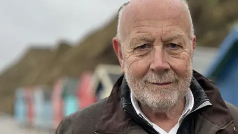Harry Blathwayt is standing in front of a row of beach huts. He has short grey hair and beard, and is wearing a white shirt, grey jumper, and a brown coat. 
