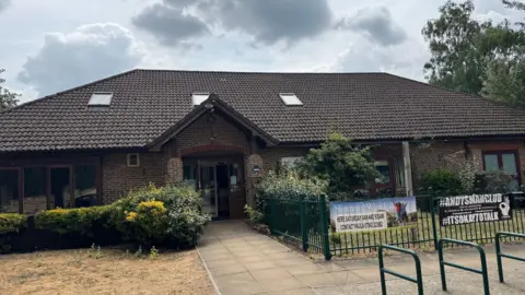 The exterior of Parkview Community Centre in Sheerwater. A one storey brown brick building with a hedge and plants in front, as well as a green metal fence.