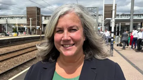 Ben Schofield/BBC Heidi Alexander smiles at the camera, dressed in a green shirt and black jacket. She is standing outside on a platform at a train station. There are a number of people in the background.