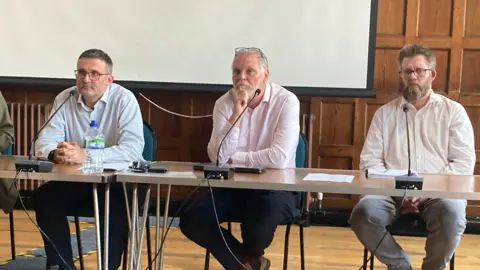 Three men wearing long-sleeved shirts sit behind a table in a public building while being questioned by members of the public about unpleasant smells at Lower Compton in Wiltshire
