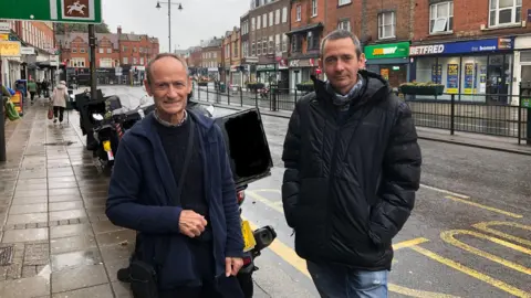 Two Councillors in front of a row of parked motorcycles on Epsom high street, which is seen in the background including shops Subway and Betfred.