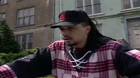 Photograph of Chris Jenkins from 1996. He is pictured wearing a black cap, a checkered red and white hoodie with a cross stitched collar. Behind him, a row of houses can be seen. 