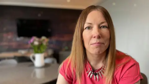 A photo of Karen looking directly at the camera. She is sitting in a home with a table behind her. She has long brown hair and a red t-shirt.
