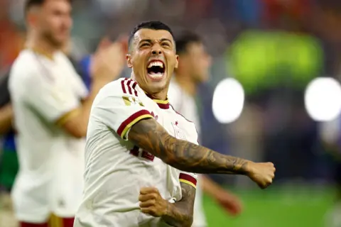 Getty Images A male footballer shouts in joy while flinging his right arm across his chest in celebration. He is wearing a white shirt with red and yellow trim and standing in a football stadium with green grass. Other players can be seen out of focus behind him.