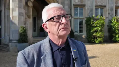 Andy Martin is standing on gravel outside Highcliffe Castle. He is wearing glasses, a dark blue shirt under a light blue jacket.