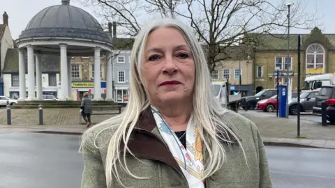 Ben Schofield/BBC Kay Mason-Billig looking direct to camera and standing in Swaffham's market square. She has straight, grey hair that reaches below her shoulders and is wearing a green, tweed overcoat and a silk scarf, that is mainly white, with patches of blue and yellow. Behind her and in the left of the shot is a rotunda with a grey, domed roof and white columns, with three steps leading up to it. Over her other shoulder is a car park, where several vehicles are parked. Further in the distance are buildings and shop fronts.