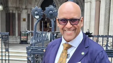 Shahrar Al Shahrar Ali stands outside the Royal Courts of Justice, wearing a dark blue blazer, white shirt, and patterned yellow tie. He has glasses and is smiling at the camera. Behind him are ornate iron gates and the stone façade of the historic court building.