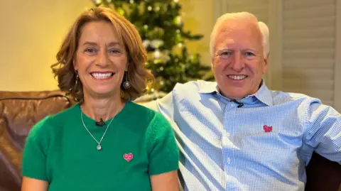 Mary Storrie wearing a green top with a pink pin badge to honour the Rosie May Foundation. Graham Storrie is sat next to her wearing a white and blue shirt, also wearing a pin badge.