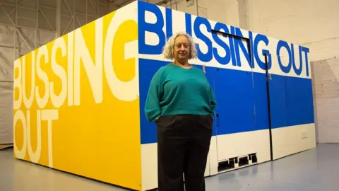Dr Shabina Aslam Lady stands in front of exhibition designed to replicate the interior of a bus.