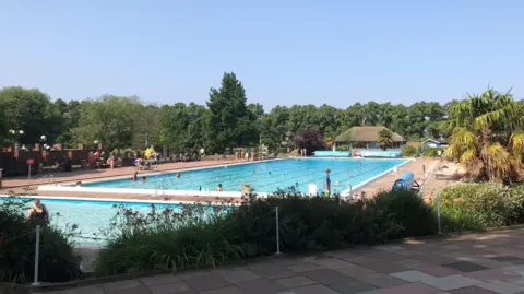 The outdoor pool seen from the far end. There is a small shallow pool and a 50-metre big pool with swimming lanes. There is a lifeguard seat on the left, with one or two lifeguards pictured wearing yellow and red uniform. There are palm trees and other trees around the sides.