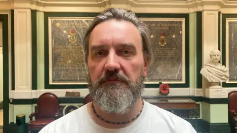 A man with grey swept-back hair and a grey and black beard wearing a white T-Shirt