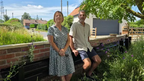 Lyn and Chris Shelton are perched on a black crash barrier which sits behind their garden wall. Lyn is wearing a green and white patterned dress whilst Chris has blue shorts and a light short-sleeved shirt on.