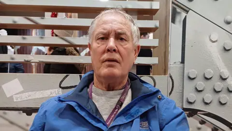 Supplied Stephen is looking at the camera with a neutral expression. He's wearing a grey jumper and blue coat, with a purple landyard around his neck.Behind him a grey industrial looking wall.