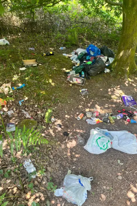 A wooded area with a small clearing that is filled with litter - there are bin bags, carrier bags and drinks cans strewn across the floor