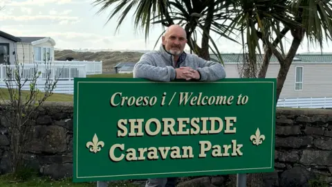 A man with a shaved head and a grey beard is leaning comfortably on top of a large green welcome sign that reads “Croeso i / Welcome to SHORESIDE Caravan Park.” He is smiling and dressed casually in a grey hoodie and dark trousers. The sign stands on a grassy area beside a stone wall, with palm-like trees and static caravans visible in the background under a bright, lightly clouded sky.