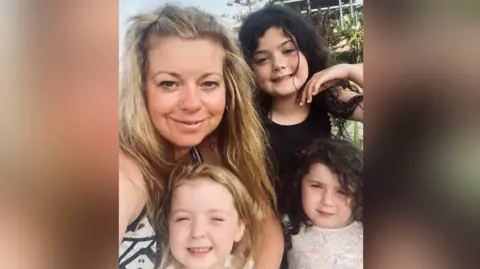 A selfie of a woman with her three children. She is on the left with blonde hair and a black and white top. There is a girl to the right of her with a blakc top nad black hair with her hand near her chin. Below her is another girl with blonde hair squinting at the camera, and next to her is a girl with a white tops with colourful dots with dark hair