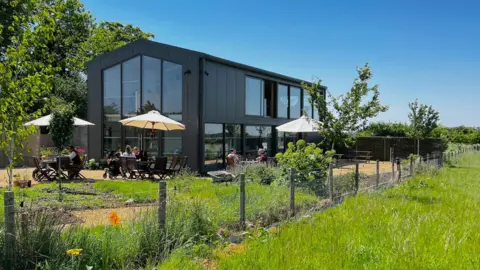 A modern building with tall windows which is used as a restaurant stands next to a grass meadow on a farm near Bath. The exterior of the building is a dark grey