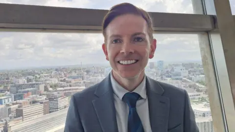 Neil Ashcroft has short hair and is wearing a grey suit, light grey shirt and blue tie. He is standing next t o a window at St Johns Beacon with a view of Liverpool city centre in the background