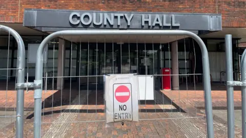 LDRS The entrance to a brick building which is blocked by fencing that has a red no entry sign attached to it. Signage on the building reads county hall in capital letters.