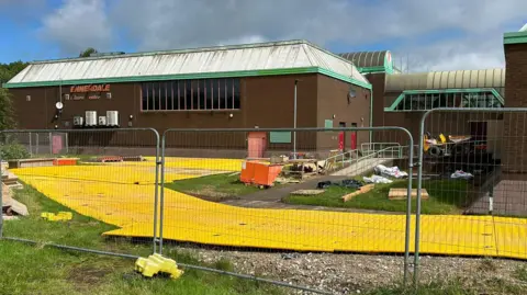 A brown brick building with green trim, displaying a sign reading “Ennerdale Leisure Centre” on the wall. In front of the building, a fenced-off area contains large interconnected yellow plastic ground protection mats spread across grass and soil. Construction materials, wooden boards, and equipment are scattered within the site. A small yellow vehicle is under a covered section to the right in the distance. Temporary metal fencing surrounds the area in the foreground, and a grassy verge runs along the edge.