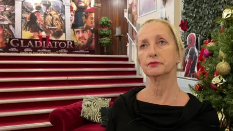 Sue Wiper has short blonde hair, and a black dress with a poppy pinned on it. On her right are some red stairs leading to a large 'Gladiator II' poster.