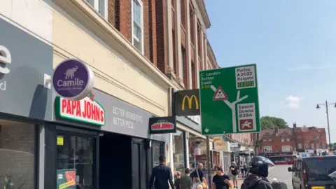 Epsom High Street. Signs for Papa Johns and McDonalds can be seen. People walk along the streets. A sign tells motorists how to get to Epsom Downs, Purley, Reigate, Central London, Sutton and Ewell.