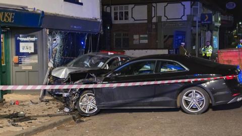 A crashed silver Ford Fiesta with a damaged parked black Audi A5 and a smashed storefront