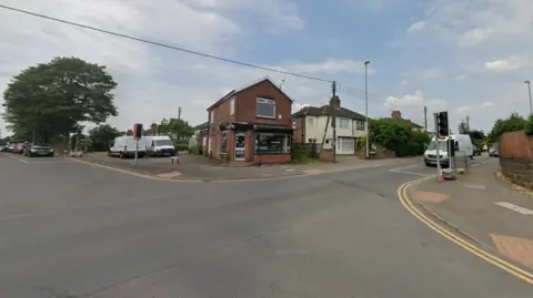 A Google street view image showing two entrances to a crossroads junction. A small shop is on the opposite side of the road, next to a white, two-storey house. Cars can be seen queuing up at traffic lights on the left and right.