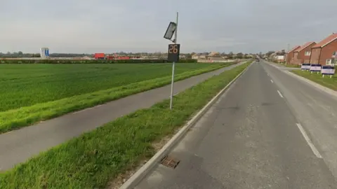 Google A road and cycle path/pavement running into a village. There is new housing on the right of the road and some construction of houses in the distance on the left. The left side of the road is a field of what looks like crops. A digital speed sign says 26 on it (presumably MPH)
