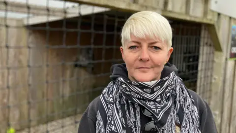 A woman stands in front of a wooden enclosure at a Bird of Prey visitor attraction.  Nets form the walls of the enclosure, which contains two Great Grey Owls. One owl is just visible in the background, sitting on a perch. The woman is wearing a patterned black and white scarf.