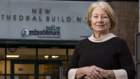 A blond woman with short hair, pearl earrings and a fine silver watch on. She is wearing a black top with a cream neck line and sleeves. Her hands are crossed and she is standing in front of a white building which has 'police ombudsman' written in blue on the entrance. 