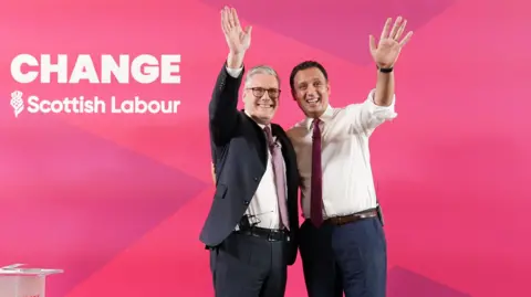 PA Media Sir Keir Starmer, left, wearing a dark suit over a white shirt and a pink tie. Anas Sarwar, right, wearing a white shirt with a red tie and dark blue trousers. Both are waving while standing in front of a red background.