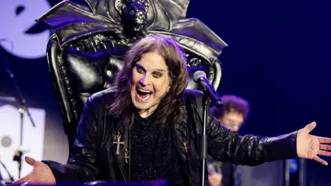 Ozzy Osbourne sat on a black throne on stage holding a microphone and looking to the audience with a wild facial expression
