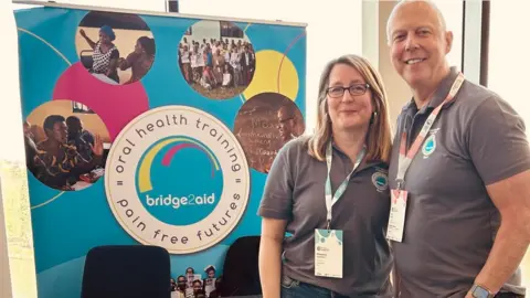 Shaenna and Phil are standing in front of a conference-style table with a large banner behind saying 'oral health training, pain-free futures and bridge2aid'. They are both wearing regulation grey polo shirts with the charity logo on and lanyards. They are smiling to camera