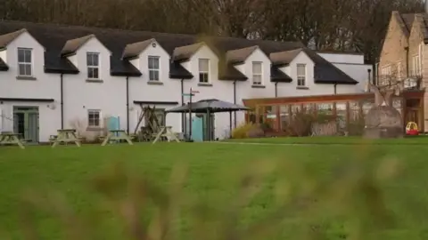 Picnic tables can be seen in the hospice garden, which has white painted walls and a veranda area overlooking the garden. A number of shrubs and plants as well as a children's plastic car can be seen in the garden.