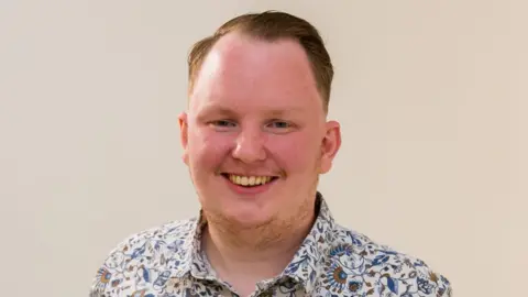 Unbranded Digital Lee Hart - a man in with short blonde hair smiles at the camera. He is wearing a floral blue and white shirt and standing against a white background