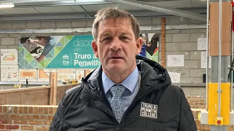 Martin has greying hair and wears a blue shirt, floral tie, and a dark raincoat. He stands inside the construction workshop with brick walls behind him.
