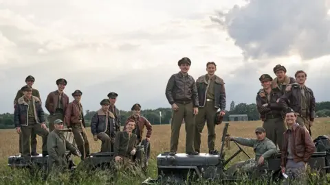 Robert Viglasky/ Apple TV Masters of the Air publicity shot showing 16 airmen in World War Two uniforms and standing on military trucks in a field.