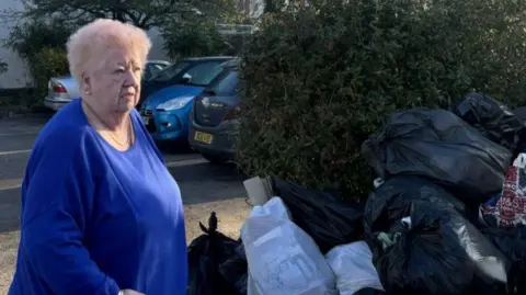 A woman with blonde hair and a blue top standing in the street in front of bin bags. She wears a blue top with a gold necklace. Behind her are several parked cars. Next to her are several bin bags in front of a hedge.