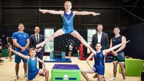 Sport NI Two young gymnasts in blue leotards kneel on the ground. Another is jumping in a star jump shape. Luke Carson and Rhys McClenaghan, in shorts and t-shirts stand behind. Gordon Lyons and Richard Archibald, in grey suits, also stand behind.