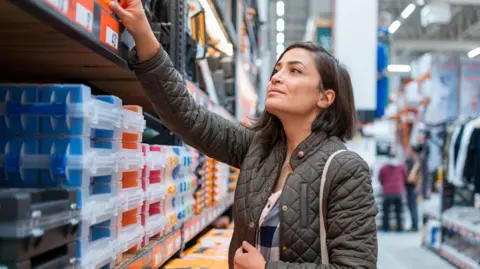 Getty images with a female dark brown hair, wear a green coat, shop in a construction store. She is reaching a shelve with boxes.