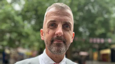 Emma Baugh/BBC Christian O'Brien smiles at the camera as he is photographed outside. He is wearing a grey waistcoat and shirt and tie. He has a beard and grey hair. He has a small graze beneath his right eye. 