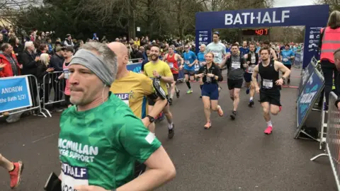 A close up of around a dozen runners with lots of spectators on the roadside and a large banner spanning the street in the background saying 'Bathalf' and a digital clock