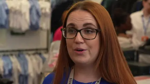 A woman with long dark red hair and glasses standing in a shop surrounded by racks of clothes. 