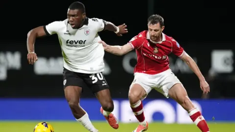 Nick Potts/PA Wire A Bristol City footballer in white t-shirt and black shorts is about to kick the yellow football as a Wrexham player in red t-shirt and white shorts starts to tackle as he grabs the Bristol players t-shirt