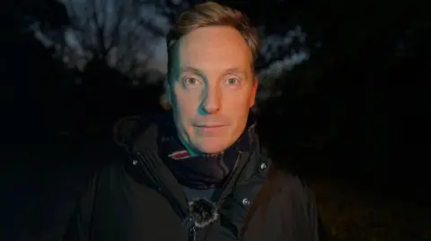 Man standing in dark. he is wearing a warm jacket and scarf and looks directly at camera.