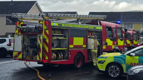 A fire appliance is parked in the middle of the road as a firefighter gathers items from an open area near the back of the vehicle. The front od a rapid response ambulance is visible in a parked area beside a number of cars.