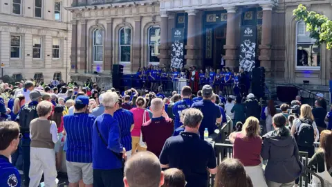 Ipswich Town Women are lined-up outside the entrance to the town hall as hundreds of fans, many dressed in blue shirts, stand in the square in front of the building.