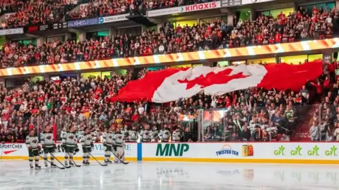 he Canadian flag is held up over spectators as the national anthem is sung prior to the start of a game between the Minnesota Wild and the Ottawa Senators at the Canadian Tire Centre in Ottowa. Ice hockey players are lined up on the rink holding their sticks. 